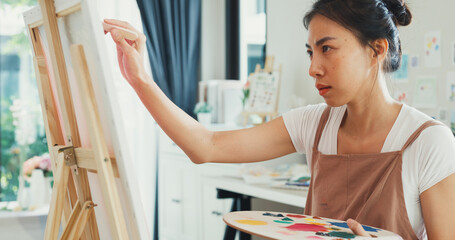 Obraz premium Close-up youth Asia lady wear apron holding palette color use brush painting on canvas create artwork in cozy workshop at home. Contemporary Painter Abstract Modern Art, Creativity and people concept.
