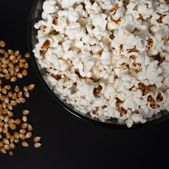 Ready-to-eat popcorn in a glass bowl and corn kernels on a black background.
