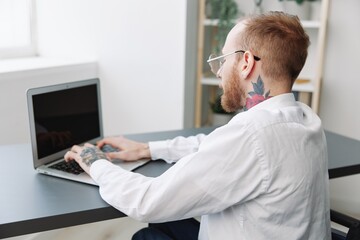 Fototapeta premium man wheelchair businessman with tattoos in the office works at a laptop online, business process from the back, copy space, integration into society, the concept of working a person with disabilities