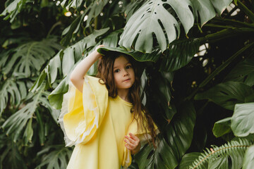 Little girl in a yellow dress in a botanical garden. a child stands near the leaves of Monstera.