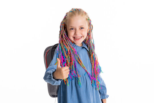 Delightful small pupil student with colorful braids shows thumb up holds painting brushes in hand wears stylish clothes and grey backpack ready to go to school pretty girl in good mood.