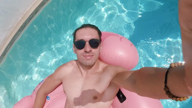 Happy man in sunglasses enjoying summer vacation filming himself swimming on a pink flamingo in a swimming pool during summer time