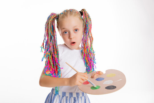 Adorable delighted schoolgirl with colorful braids holding painted tools shows surprised emotions standing on white background isolated.