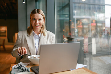 Smiling businesswoman is having a business lunch and working on laptop in cafe. Blurred background