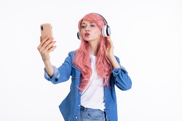 Young woman mother wearing pink wig and modern headphones holding smartphone in hand speaking by video link sending kisses at screen while posing on white background in studio isolated.