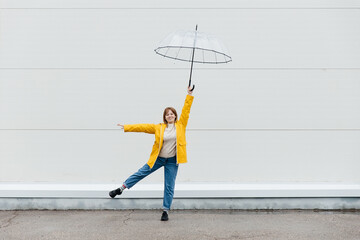 Young girl dressed in a yellow raincoat holds a transparent umbrella and poses near a white wall