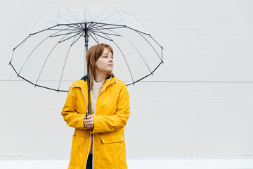 Young girl dressed in a yellow raincoat holds a transparent umbrella and poses near a white wall