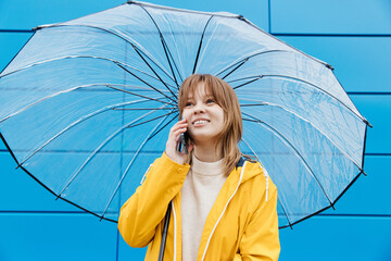 Young girl dressed in a yellow raincoat with an umbrella and a smartphone against a blue wall