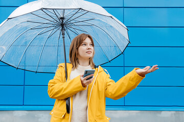 Young girl dressed in a yellow raincoat with an umbrella and a smartphone against a blue wall