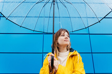 Woman dressed in a yellow raincoat with a transparent umbrella against a blue wall