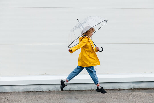Young girl dressed in a yellow raincoat holds a transparent umbrella runs near a white wall