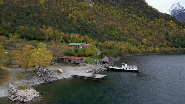 Drone flies between old boathouse and sawmill on Vike in Eikesdsal, in Molde municipality in Norway. The old ferry is moored at the quay. Beautiful autumn colors characterize nature on this day.