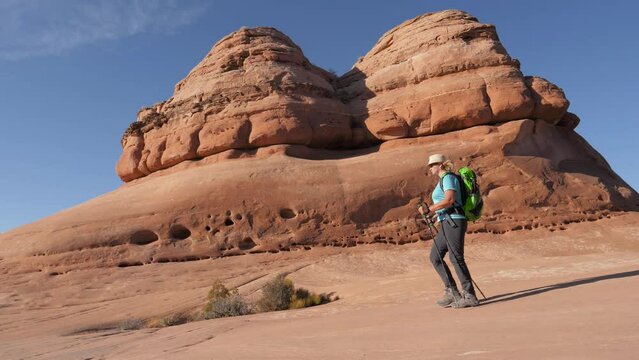 Hiker mature woman with backpack and trekking poles hiking at flat and smooth rock monolith of orange sandstone. Walking background rock formations in Arches national park Utah. Slow motion side view
