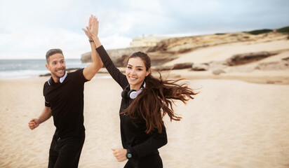 Happy man and woman in sportswear giving five while running together, training by seaside, panorama, free space
