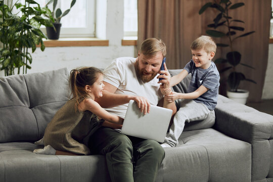 Man, father sitting on couch, talking on phone and working on laptop in living room. Kids bothering and playing with him. Concept of fatherhood, childhood, family, freelance job, remote work