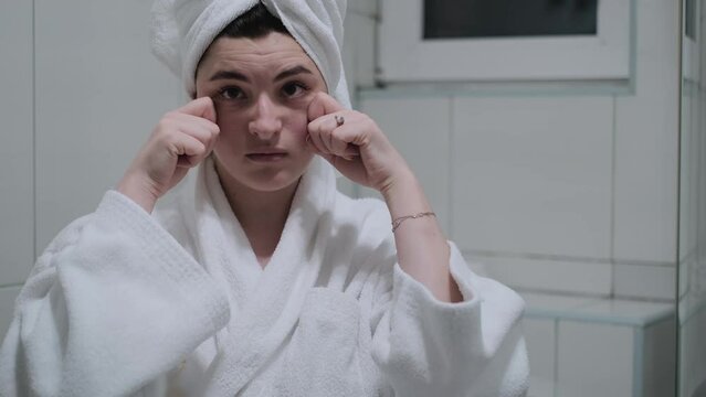 A woman in a bathrobe looks in the mirror and gives herself a facial massage. Touch the skin with your fingertips, enjoy an easy effective anti-wrinkle treatment at home. Removal and prevention of mim