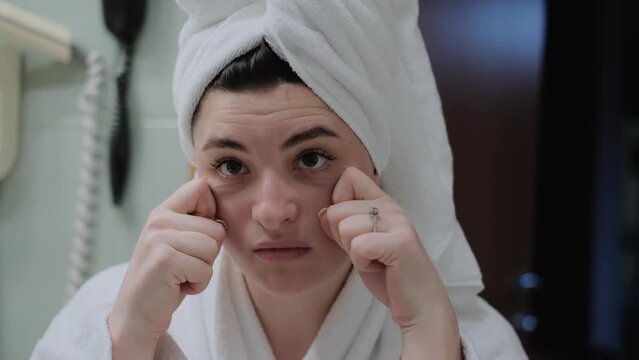 A woman in a bathrobe looks in the mirror and gives herself a facial massage. Touch the skin with your fingertips, enjoy an easy effective anti-wrinkle treatment at home. Removal and prevention of mim