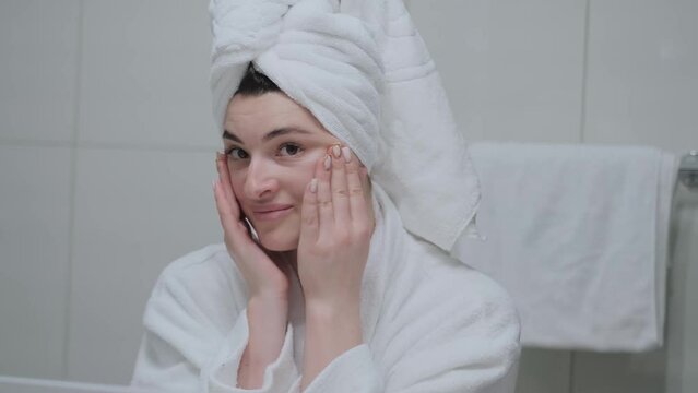 A woman in a bathrobe looks in the mirror and gives herself a facial massage. Touch the skin with your fingertips, enjoy an easy effective anti-wrinkle treatment at home. Removal and prevention of mim