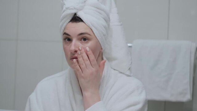 A woman in a bathrobe looks in the mirror and gives herself a facial massage. Touch the skin with your fingertips, enjoy an easy effective anti-wrinkle treatment at home. Removal and prevention of mim