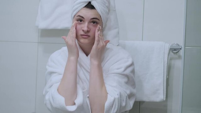 A woman in a bathrobe looks in the mirror and gives herself a facial massage. Touch the skin with your fingertips, enjoy an easy effective anti-wrinkle treatment at home. Removal and prevention of mim