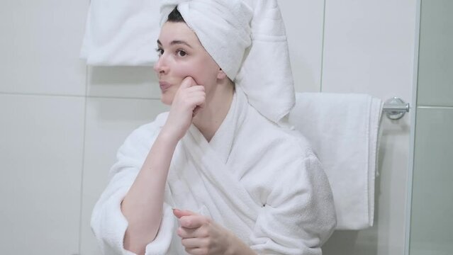 A woman in a bathrobe looks in the mirror and gives herself a facial massage. Touch the skin with your fingertips, enjoy an easy effective anti-wrinkle treatment at home. Removal and prevention of mim