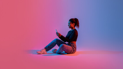 Full length of black lady sitting with cellphone, surfing social media or chatting in colorful neon...