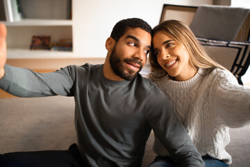 Smiling pretty millennial european female and arab guy have fun, take selfie together in living room interior