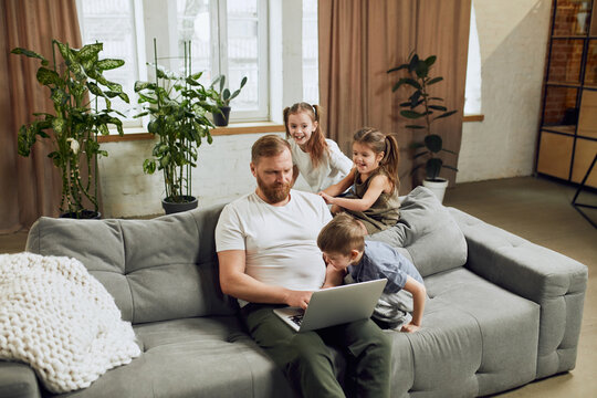 Mature man, father sitting on couch at home and working remotely on laptop with three little kids playing around him and making noise. Fatherhood, childhood, family, freelance job, home office concept
