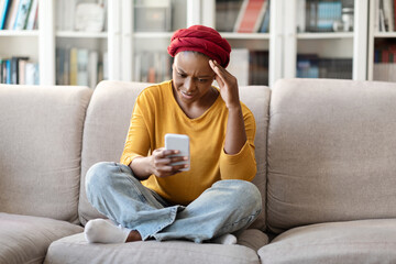 Upset black woman sitting on sofa, using phone, home interior
