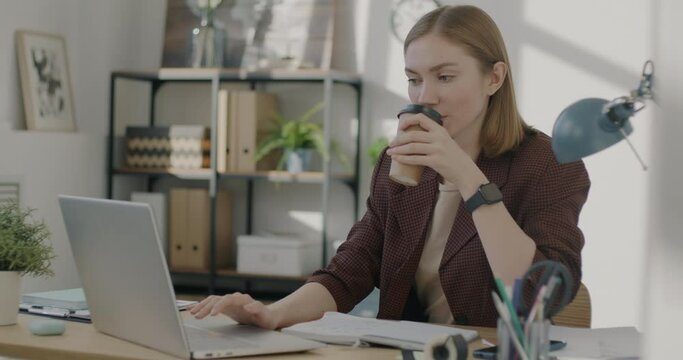 Female employee working in office with laptop and drinking take out coffee from single use cup in modern office. Job and beverage concept.