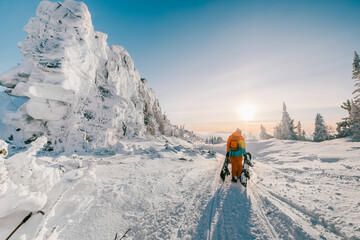 Snowboarder walking with a snowboard between beautiful rocks covered with snow and trees. Ski touring in the snowy mountains during sunset