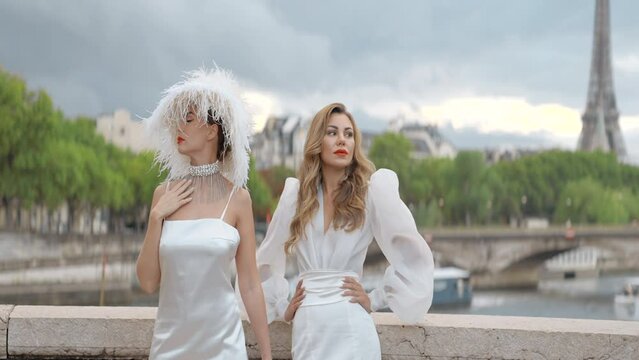 Stylish models in white dresses posing on the background of Paris streets and Effel tower. Action. Women in elegant dresses outdoors, one of them wearing a hat with feathers.