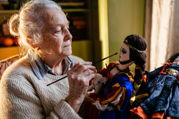 Old Caucasian beautiful grey haired artist woman sewing doll at home. Maker exclusive colorful handmade needlework.