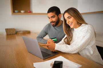 Smiling millennial arab guy and european female pay for order on laptop, use credit card, sit at table in room