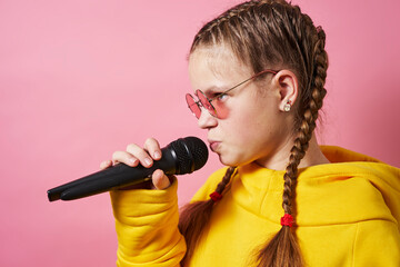A girl in a yellow hoodie and pink glasses holds a microphone and emotionally performs a song....