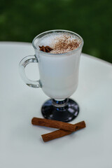 A tall glass with coffee latte and cinnamon sticks stands on a white table with a garden in the background