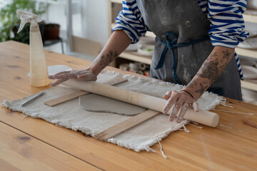 Hands of artisan woman processing clay with wooden rolling pin to make original handmade crockery. Close-up of process of creating craft souvenirs by girl in dirty apron with tattoos on wrists