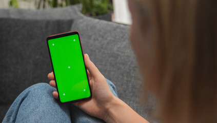 Back view of young europen woman who is using chroma key smartphone screen.She is sitting on the sofa in her cozy living room.