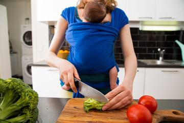 Woman with baby in a wrap carrier cooking in the kitchen, cutting vegetables with knife on a chopping board. Interior of a cozy modern kitchen. Mother doing her chores. 
