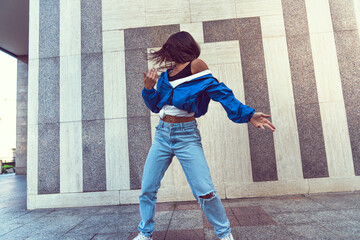 Photo shoot of stylish caucasian hip hop dancer posing having fun in blue jacket, jeans, black top and white tshirt, enjoy the urban town as background. City buildings shooting of moving fitness model