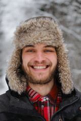 Happy emotional guy on walk in park portrait close-up. Caucasian bearded young handsome man wears fluffy hat with earflaps and stands in winter forest during snowfall.