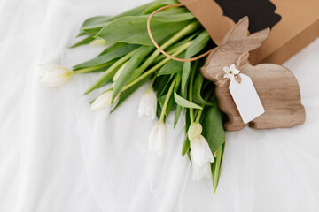 The Wooden Easter Bunny sits near a gift bag with white tulips on a light white background. Spring Background for Easter. International Women's Day, March 8th