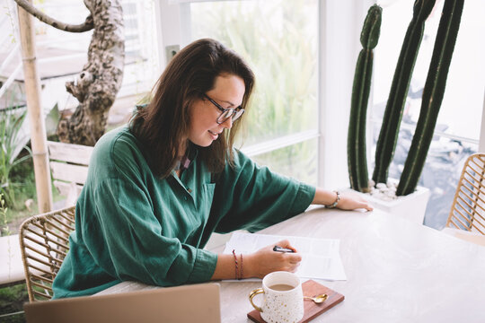 Intelligent business woman in glasses analyzing report documents during free time in cafeteria, Caucasian female employee in optical eyewear editing paperwork while doing course work in cafe interior