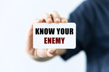 Know your enemy text on blank business card being held by a woman's hand with blurred background. Business concept about knowing opponents.