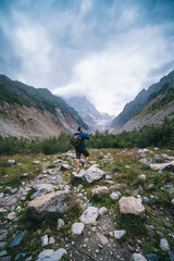 Fototapeta na wymiar Traveler man with a backpack standing on a stone, On the way, walk the track. A valley among majestic high mountains.. Back view. The Chalaadi Glacier, Georgia, Caucasus. Vertical photo.