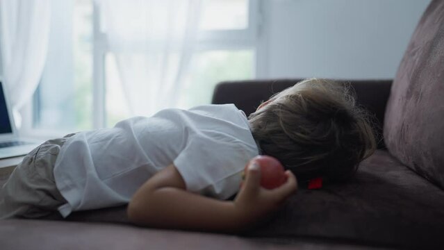 Child lying on couch with apple fruit. One male caucasian kid eating healthy snack laid on sofa at home. Healthy nutritious diet