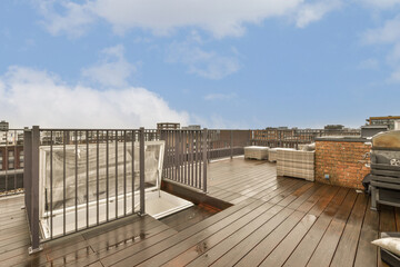a rooftop with wood flooring and metal railings on the roof, against a blue sky filled with white...