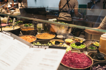 Person browsing the menu card in front of the counter of a vegan and health food restaurant filled with bowls of food.