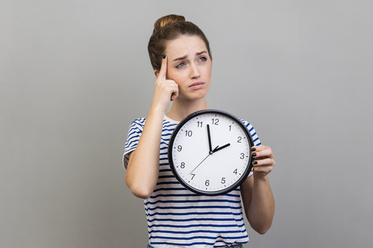 Pensive thoughtful woman wearing striped T-shirt holding wall clock, thinking about deadline.