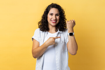 Woman standing, looking at camera, pointing at her smart watch, expressing happiness.